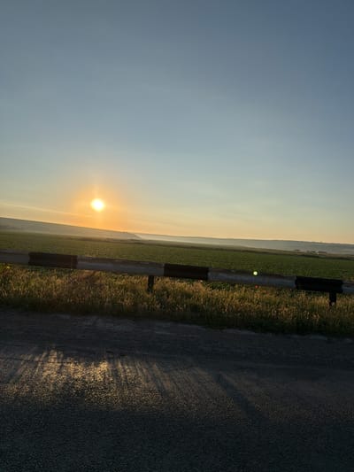 This image depicts a sunset over a rural landscape in Cîrnățeni, Moldova. The sun is a bright orb low in the sky, casting a warm glow across a vast field of green crops. A guardrail runs horizontally across the middle of the frame, separating the field from a dark asphalt road in the foreground. The road surface reflects the sunlight in a pattern of long shadows. The sky transitions from a pale blue at the top to a soft orange near the horizon, with a few wispy clouds visible. The overall scene conveys a sense of peace and natural beauty.