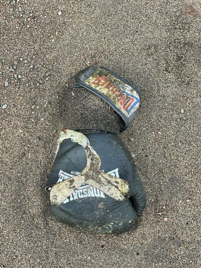 The image shows a damaged black boxing glove lying on a sandy beach. The glove is partially covered in sand and what appears to be seaweed or other organic debris. The brand name "Lonsdale" is visible on the glove. A separate strap from the glove, also dirty and worn, lies near the main body of the glove. The beach appears to be in Dauin, Philippines, given the context. The scene depicts a piece of discarded sports equipment on a natural beach environment. There are no people or animals visible in the image.
