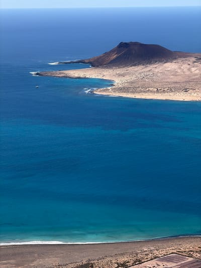 This aerial photograph captures a dramatic and arid coastline under a clear, bright sky. If viewed with the landscape oriented horizontally, the left side is dominated by a rugged, dark, conical mountain or volcanic peak. This landmass slopes down, forming several distinct rocky headlands that jut into the deep blue ocean.

To the right, a long stretch of sandy and pebbly beach forms a clear boundary with the water, where gentle waves can be seen breaking into white foam. Beyond the beach, the land continues its arid character, featuring sparse vegetation and a visible pattern of rectangular plots or structures, possibly related to agriculture or human development, further inland.

The ocean itself is a vibrant blue, ranging from a darker, deeper hue offshore to a lighter, turquoise shade closer to the coast, indicating varying depths. Several small boats are visible as tiny specks in the water, particularly near the rocky protrusions on the left, suggesting maritime activity.

The time of day appears to be mid-day, with strong, clear lighting and no clouds in the sky, indicative of excellent weather. The barren, volcanic terrain, and the intense blue of the ocean are strong geographical cues consistent with the Canary Islands, where Haría, Spain, is located. The scene evokes a sense of natural grandeur and tranquility. There is no visible text in the image.