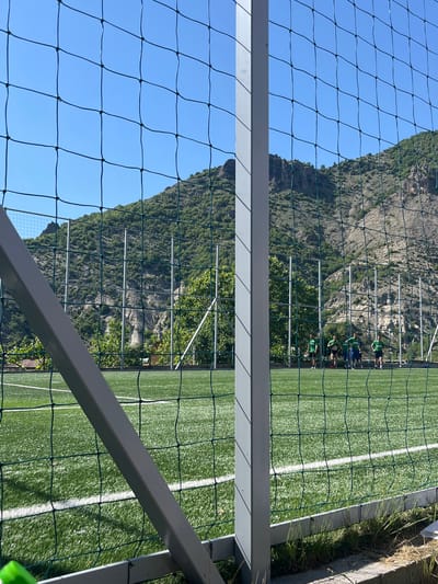 A view through a fence and a metal structure of a soccer field in Bukovo, Bulgaria. The field has artificial turf and white lines. A group of people wearing green shirts are visible along the field. In the background, a mountain rises behind the field against a clear blue sky. Grass and concrete border the field on the right side of the image.