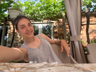 The image shows a young woman sitting on a wicker chair outdoors, with a table in the foreground. She is wearing a grey tank top and a silver necklace. Behind her, there is a brick wall, a window, and green foliage from a grapevine. To the right, there is a sheer curtain and another wicker chair. The scene appears to be in a garden or patio area. The specific location in Rogozen, Bulgaria, is not identifiable from the visual information provided.