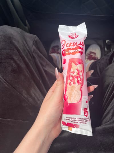 A person is holding a pink and white popsicle wrapper with an ice cream image on it. The wrapper has Russian text. The person's hand, with long, light-colored acrylic nails, is holding the wrapper. The person is wearing dark, loose-fitting pants. The background shows a dark interior, possibly a car, with blurred feet wearing white shoes with red accents visible. The location is identified as Novaya Otradovka, Russia.