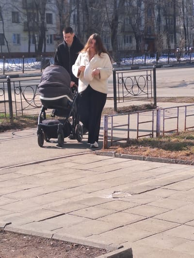 A man and a woman are walking on a concrete slab sidewalk in the city of Меткомелино, Russia. The adult male, dressed in a dark hooded jacket and dark pants, pushes a dark gray baby stroller with his hands on the handle. The adult female, with long brown hair and wearing a light-colored jacket over dark pants, walks to the right of the stroller, looking down at a light-colored object held in her hands. The sidewalk consists of uneven and cracked square concrete paving slabs. To the immediate left of the sidewalk, a decorative black metal railing separates it from an asphalt street. Further beyond this railing, a low, faded lavender-painted metal barrier borders a strip of ground with dry, brown grass and patches of melting snow. The background features numerous deciduous trees with bare branches, partially obscuring multi-story residential buildings. The scene is illuminated by bright sunlight, indicative of a clear day.