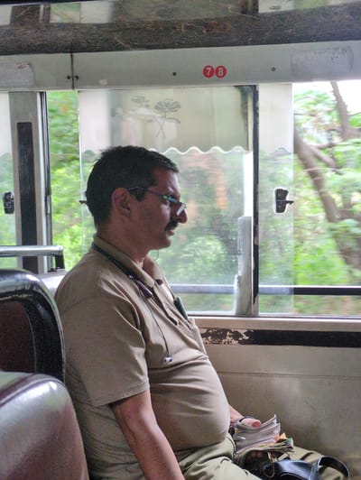 The image shows a man, possibly a bus conductor, sleeping on a bus. He is wearing a tan uniform, a shirt with a collar, and tan trousers. He has glasses on and his head is tilted back, with his mouth slightly open. A few items are visible on his person, including a pen clipped to his shirt pocket, and a small black strap attached to his collar. He appears to be holding a clear plastic case with some papers or tickets inside. The bus interior is visible, with a window showing green foliage outside, suggesting a rural or semi-rural setting, perhaps during the day. The scene appears to be taking place in Bajaura, India. On the side of the bus, a red circular sticker with the number "78" is visible. The overall mood seems relaxed, with the man resting during his duty.