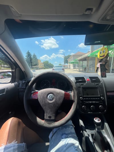 The image, taken from the interior of a car, shows a view looking forward and slightly to the right through the windshield. The car's steering wheel, with a Volkswagen logo, is prominently in the foreground. The dashboard features an infotainment screen, air vents, and controls. A manual gear shifter is visible to the right of the steering wheel. Outside the car, a street scene unfolds with buildings, trees, and other vehicles. The sky is blue with scattered clouds. Based on visual cues such as the architecture of the buildings and the signage on the awnings, the location appears to be the town of Ботево, Bulgaria. The scene is set during the daytime under clear skies.