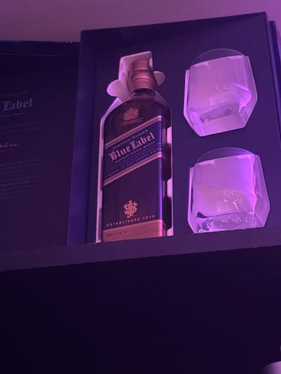 The image shows a Johnnie Walker Blue Label whisky set inside a dark box. The set includes the bottle of whisky and two drinking glasses. The box appears to be black. The lighting is poor with a purple hue. The image does not depict the city of Mamer, Luxembourg and does not include any essential details about people, actions, or interactions.