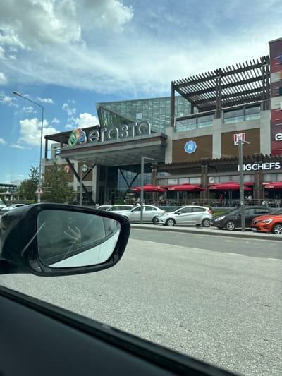 The image captures a view from inside a vehicle, with a side mirror in the foreground. Reflected in the side mirror is part of the facade of Erasta Edirne, a shopping center in Edirne Merkez, Turkey. The shopping center features a glass and steel structure with signage displaying the "Erasta" logo. The architecture includes pergolas and outdoor dining areas with red umbrellas. Adjacent to Erasta Edirne, there are branded restaurants, including KFC and BigChefs. Several parked cars line the street. The sky is visible with scattered clouds.