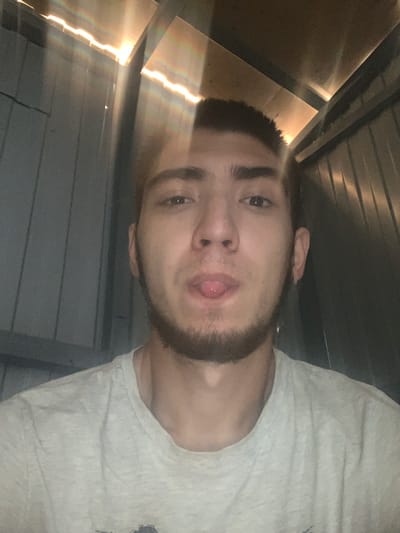 A young man with dark hair, a short beard, and a light-colored t-shirt is pictured in a selfie. He is sticking his tongue out at the camera. The background consists of corrugated metal walls, with light streaming in from above, creating a rainbow effect. The image appears to have been taken indoors, possibly in a shed or storage unit. There are no discernible landmarks or environmental clues to indicate the specific location within Kokhma, Russia.