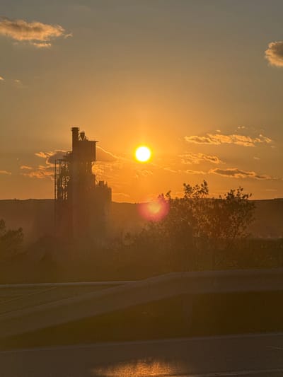 The image depicts a sunset in Kipra, Bulgaria. The sun is positioned high in the sky, radiating a bright orange glow that casts long shadows across the landscape. A silhouette of a building or structure is visible against the bright sky. The sky has some patchy clouds, illuminated by the sun. There is a dark shadow to the right with blurred objects in the foreground.