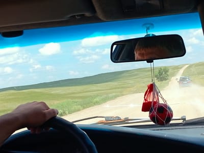 The image shows a view from inside a vehicle, likely in the city of Aleksandrovka, Kazakhstan, as indicated by the terrain. A hand is visible on the steering wheel. A rear-view mirror reflects the head of a person and the sky. Hanging from the mirror are a red bag and a red and black soccer ball in a net. Outside the vehicle, a dirt road stretches into the distance, flanked by grassy hills. Two vehicles are visible on the road ahead, with dust trails indicating their movement. The sky is blue with scattered white clouds.