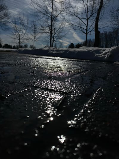 This image depicts a low-angle view of a thawing winter or early spring scene in Novoivanovskoe, Russia. The foreground features a dark, wet paved surface, likely a sidewalk or pathway, consisting of rectangular or square units. Numerous points of bright sunlight are reflected on the wet surface, indicating the presence of water and ice, with some reflections exhibiting subtle purple and pink hues. Shallow puddles and streaks of water are visible across the dark paving.

In the midground, a distinct bank of white snow with an irregular, melting surface runs horizontally, separating the paved area from the background.

The background presents several bare deciduous trees with intricate branch structures silhouetted against a partly cloudy sky. The sky shows varying shades of blue and grey, with prominent white contrails or cirrus clouds stretching across it. Beyond the initial line of bare trees, a denser, darker mass of trees suggests a wooded area or forest. No individuals, vehicles, or specific architectural structures are discernible. The scene is illuminated by strong, low-angle sunlight, casting reflections and silhouetting elements, consistent with late winter or early spring conditions.