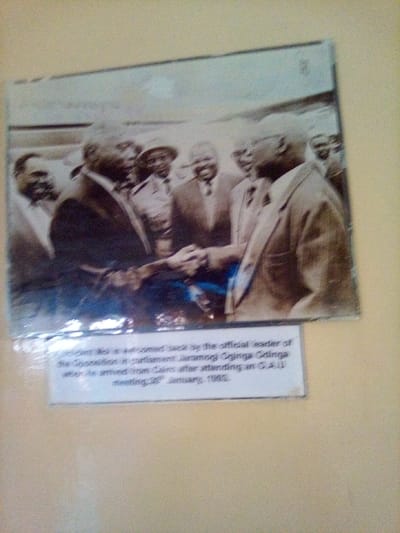 A framed sepia-toned photograph with an accompanying white caption label is mounted on a light-colored wall in the city of South Sakwa ward, Kenya. The photograph depicts approximately eight adult males in formal attire, centered on a handshake between two prominent figures. The man on the left, identified by the caption as President Moi, wears a dark suit, facing right and extending his right hand. The man on the right, identified as Jaramogi Oginga Odinga, wears a lighter suit, facing left and extending his left hand. Other men are visible in the background, observing the handshake; one man in the mid-left has a beard, and another in the background wears a hat. The caption beneath the photo reads: "President Moi is welcomed back by the official leader of the Opposition in parliament Jaramogi Oginga Odinga when he arrived from Cairo after attending an O.A.U meeting, 30th January, 1983." The display exhibits some surface glare.