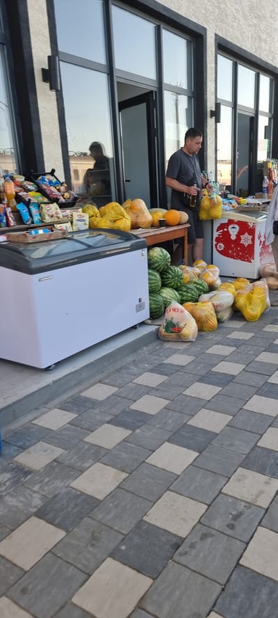 The image shows an outdoor market stall in Şïeli, Kazakhstan, with a man standing next to a display of produce. The stall features a large white chest freezer, a wooden table laden with pumpkins, melons, and watermelons, and shelves stocked with snacks and bottled drinks. In the background, the entrance to a building with large glass windows is visible, reflecting the sky and passersby. The ground is paved with interlocking gray and beige tiles.