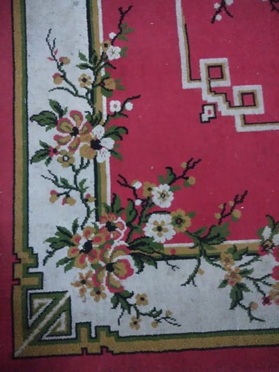 The image displays a close-up, top-down view of a red patterned rug or carpet. The design prominently features a large central field of off-white, bordered by parallel thin stripes of gold and olive green. Within this off-white field, a dense floral pattern is present, consisting of green vines, leaves, and various flowers rendered in white, yellow, and pink with dark centers. In the upper-right portion, against the red background, a distinct white and gold-outlined geometric meander pattern is visible. The lower-left corner of the off-white field incorporates a more elaborate geometric motif with diagonal green and white elements. The textile exhibits a visible woven texture with individual fibers and minor surface dust accumulation, particularly on lighter areas. No individuals, additional objects, actions, interactions, or discernible environmental context are present beyond the carpet itself. The specific location within the city of Chimay, Belgium, cannot be ascertained from the visual evidence provided.
