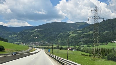 This is a photograph taken from a moving vehicle on a highway in St. Georgen ob Judenburg, Austria. The highway curves to the right, with a concrete barrier on the left and a metal guardrail on the right. Several cars are visible on the highway. The landscape features rolling green hills covered in dense forests, with scattered houses and buildings on the slopes. A small town or village is nestled in a valley to the left of the highway. A large electricity pylon stands prominently in the foreground on the right side of the image. The sky is blue with scattered white clouds. The image captures a scenic view of the Austrian countryside along a major roadway.