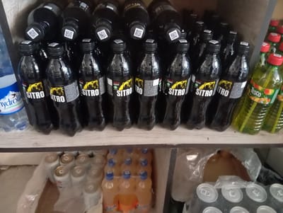 A close-up, slightly blurry, eye-level shot shows a shelf filled with drinks. On the top shelf, there are rows of black bottles with yellow and white labels that read "SITRO". To the right of these bottles are several green bottles with red caps and yellow labels. A portion of a clear bottle with a blue label is visible on the far left. The bottom shelf contains rows of canned beverages wrapped in plastic. To the right of the cans are several orange-colored bottles with blue caps, also wrapped in plastic. The background is a rough, greyish surface. The image appears to be taken in a retail setting in Yangikurgan, Uzbekistan.