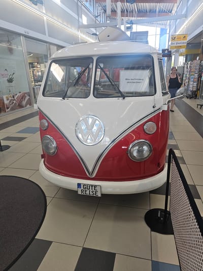 This is a vertical, medium-shot photograph of the front of a classic red and white Volkswagen Type 2 Kombi van displayed indoors. The van has a prominent chrome Volkswagen logo on its front. A German license plate with the text "GUTE REISE" (meaning "Have a good trip") is visible on the lower right of the van. A white sign with black text is placed on the windshield of the van. A woman wearing a black top and blue shorts is standing behind the van, looking towards the right of the frame.

The van is parked on a tiled floor in what appears to be a public indoor space, possibly a shopping mall or a terminal, given the presence of various shops and signage in the background. Signs for "Postbank," "Der Postladen," and "LOTTO" are visible, suggesting a service area. The lighting suggests it is daytime. The overall scene is clean and well-lit, with the classic vehicle being the clear focal point. The woman's expression is neutral. The van is in pristine condition, indicating it's likely part of an exhibition or display.