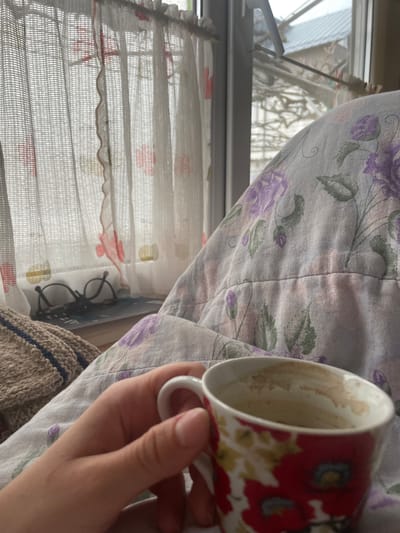 A person holds an empty, floral-patterned mug in their right hand, next to a light-colored floral-patterned fabric, likely a blanket or bedding. Behind the hand and blanket is a window with a white, semi-transparent curtain. Through the window, there are blurry buildings and structures. Near the bottom of the frame, glasses are visible under the curtain, near the edge of a brown knitted blanket. The location is likely indoors, possibly Aprilovo, Bulgaria. The interaction involves a person holding an empty mug indoors next to a window.