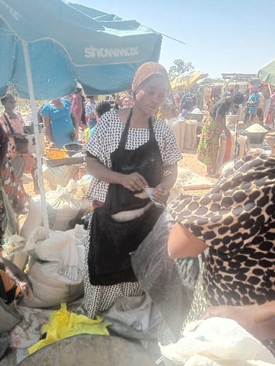 This image depicts an outdoor market scene in Gurku Gwari, Nigeria. In the foreground, a woman wearing a white patterned dress, a black apron, and a brown head covering stands under a blue umbrella bearing the "showmax" logo. She is holding and appearing to clean or scale two fish. To her right, another individual is partially visible, wearing a leopard-print top.
The ground is dusty and covered with numerous white and light-colored sacks, some containing grains or other market goods. A yellow plastic bag is visible on the ground near the sacks. In the background, multiple individuals are present, engaging in various market activities. One woman in a blue dress stands near some yellow containers. Further back, more market stalls with umbrellas and various goods, including additional sacks, are visible under a bright, clear sky, suggesting daytime conditions. The overall scene indicates a busy, bustling market environment.