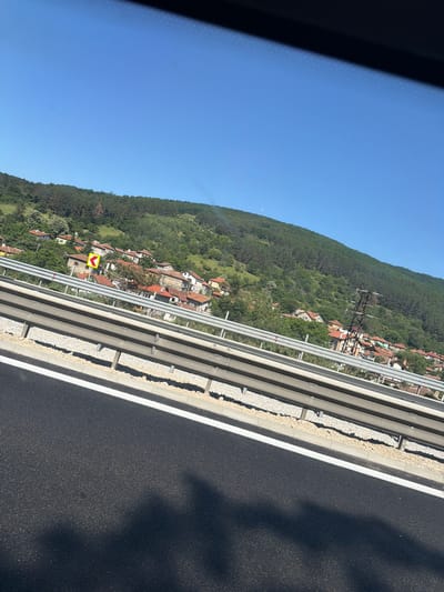 The image captures a view from a vehicle traveling on a road, likely a highway, overlooking a village nestled on a hillside. The sky is a clear, bright blue. In the foreground, the dark asphalt of the road is visible, marked with a white line. A metal guardrail separates the road from the area beyond.
Beyond the guardrail, a cluster of houses with reddish-brown roofs are spread across the green, tree-covered slope of a hill. The architecture appears to be typical of a rural European village. A yellow road sign with a black arrow indicating a curve to the left is visible near the houses. Further up the hill, dense green forest covers the landscape, transitioning into a darker, more rugged terrain at the top. A tall, lattice-style electricity pylon stands prominently on the hillside, a testament to the infrastructure supporting the area. The overall impression is of a sunny day in a scenic, semi-rural setting, potentially near the town of Churek, Bulgaria, given the context provided. No people or specific actions are discernible in the image.