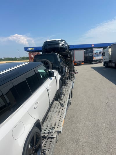 In St. Marienkirchen bei Schärding, Austria, a car carrier is parked on an asphalt surface, likely at a service area or truck stop. The carrier is loaded with several vehicles, including two white Land Rover Defenders in the foreground and a black one stacked on top. In the background, a blue structure with the "AS24" logo is visible, suggesting a fuel or service station. Several trucks are also parked nearby, with one bearing the "INTERCOM GROUP" logo. The sky is clear with some scattered clouds, indicating a sunny day. The overall scene depicts a logistics operation involving the transport of vehicles.