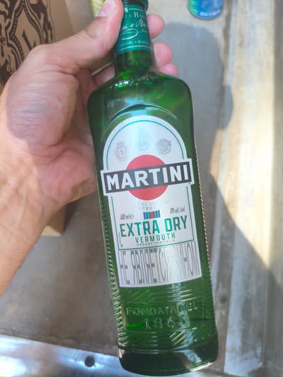 The image shows a hand holding a green glass bottle of Martini Extra Dry Vermouth. The bottle has a white label with the Martini logo, the text "MARTINI", "EXTRA DRY", "VERMOUTH", "500 mle", and "18% alc./vol.". The bottle also has the text "FONDATA NEL 1863" embossed on the glass near the bottom. The background is out of focus and appears to be a concrete surface and some fabric. There is no indication of the location being Alchevsk, Ukraine.