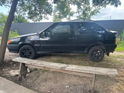 A black, three-door hatchback, possibly a Lada, is parked on a dirt area next to a gray metal fence. A weathered wooden bench with a rough-hewn appearance sits in front of the car. A mature tree with a textured trunk is positioned to the left of the car, with its branches and leaves casting shadows over the scene. The ground is covered in dirt, scattered leaves, and numerous cigarette butts. In the background, green foliage and a cloudy sky are visible. The location is identified as Pereiaslavske, Ukraine. There are no people visible in the image, and no apparent actions are taking place.