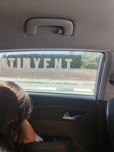 An interior view from the backseat of a car looking out the rear window at large, white, three-dimensional letters spelling "ONTINYENT". The letters are situated in a landscaped area with gravel and greenery, likely an entrance to the city. A child's head and shoulder, with long brown hair, are visible in the foreground, leaning against the car door. The car's interior, including a door handle and part of a seat, is also visible. The lighting suggests it is daytime.