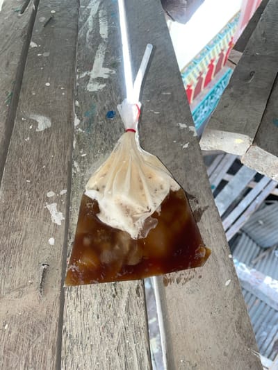 A clear plastic bag containing a dark brown liquid with lighter brown foam and translucent solid particles is depicted resting on a weathered, light gray wooden plank surface. A white plastic straw is inserted into the bag, which is secured at the top with a red elastic band. The wooden surface exhibits white, blue, and green paint splatters and visible nails. In the background, partially obscured, a building with a light blue railing and red, stylized balusters is visible. Additional indistinct structures, possibly corrugated metal, are present in the lower right background. The scene is located in Mns Asan, Indonesia.