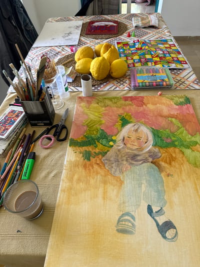 The image shows a cluttered table in what appears to be a residence, possibly in Kfar Vradim, Israel. Dominating the scene is a partially completed painting of a young girl lying down in a grassy area, rendered in watercolor and pastel hues. To the left of the painting is a game of Snakes and Ladders, crayons in a plastic container, and a bowl of lemons. Scattered around are various art supplies including paintbrushes in a cardboard box, a thermos container, watercolor paints, pencils, a highlighter, scissors, and a glass filled with liquid. Jeans are visible in the bottom left corner. The surface the items are placed on is a tablecloth with two different patterns. The scene suggests an artistic hobby and/or pastime.