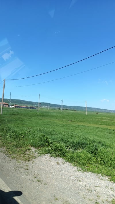 The image depicts a rural scene near Straldzha, Bulgaria. The sky is clear blue with a few wisps of clouds. A grassy field dominates the foreground, with varying shades of green suggesting different vegetation. A gravel road or shoulder is visible along the bottom edge of the frame. Utility poles and power lines stretch across the landscape, receding into the distance. In the mid-ground, some buildings and a fence line are discernible. A low mountain range forms the horizon. No people are directly visible in the shot. The picture appears to be taken from a moving vehicle.