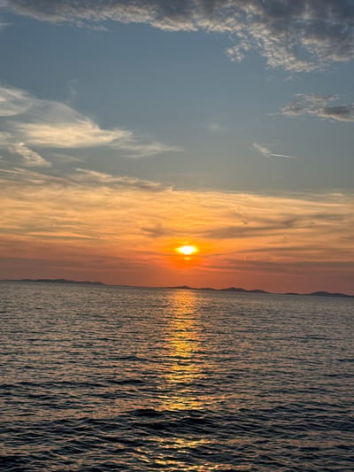 The image depicts a sunset over the Adriatic Sea, viewed from the vicinity of Primošten, Croatia. The sun, a bright orb, is setting on the horizon, casting a warm, orange glow across the sky and reflecting on the water. The sky transitions from a pale blue at the top to vibrant shades of orange and yellow closer to the horizon, with scattered wispy clouds. In the distance, a silhouetted chain of islands forms a low, undulating line against the colorful sky. The sea in the foreground is dark blue, with ripples catching the sunlight to create a shimmering path. The overall scene is serene and captures the natural beauty of a coastal sunset.