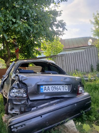 In Pishchanka, Ukraine, a severely damaged dark grey sedan is positioned outdoors. The vehicle's rear is heavily crushed, with the trunk area appearing warped and the rear window completely shattered. The license plate, UA AA 0964 TC, is visible. A separate dark grey bumper lies in front of the car, seemingly dislodged. The car is situated on a grassy area, with a rustic wooden fence and a building with a corrugated metal roof visible in the background. A boy, wearing a striped t-shirt and red shorts, is climbing a tree to the left of the car. The environment suggests an outdoor rural or semi-rural setting, possibly a backyard or a junkyard.