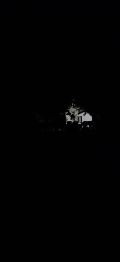 The image depicts a dark, grainy scene that appears to be a nighttime exterior shot. In the center-right portion of the frame, a white building with visible windows and a roofline is partially obscured by dark foliage, likely trees. The building is dimly lit, with some internal illumination suggested by the lighter areas within the windows. The overall impression is one of low light conditions, possibly taken in a rural or suburban setting. The specific location in Orzhiv, Ukraine, is not discernible from the visual information provided in the image.