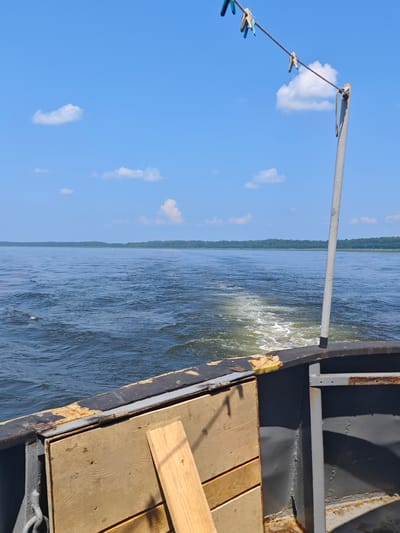 The image depicts a view from the back of a boat moving through a body of water under a clear blue sky with scattered white clouds. The water shows a visible wake and churned foam behind the boat. The foreground shows the weathered deck of the boat with some wooden planks. A clothesline with several clothespins is strung across the upper right portion of the image. In the distance, a treeline of dense green foliage borders the horizon. The scene appears to be set on a calm, sunny day, likely in a rural or natural setting. The specific location in the city of Погодаево, Russia, is not discernible from the image itself, as there are no identifiable landmarks or structures that would indicate this specific geographic area.