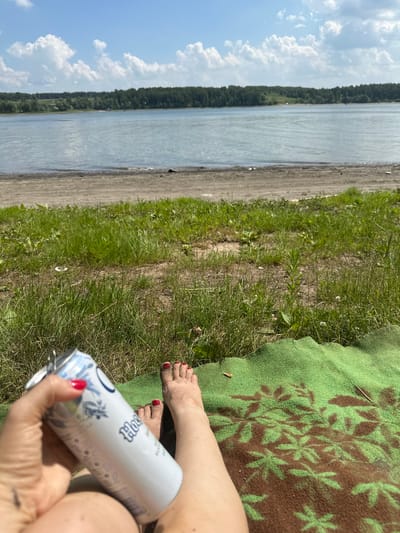 The image depicts a person's legs and hand holding a beverage can, resting on a green blanket with a brown floral pattern. The person's feet are bare and appear to have dirt on them, with bright red nail polish on the toenails. A white can with blue accents and text is held in the hand. The scene is set outdoors with a body of water in the background, a sandy shore, and lush green grass. A line of trees borders the water, and a blue sky with white clouds is visible above. This appears to be a tranquil outdoor setting, possibly a picnic or relaxation by the water on a sunny day. The specific location in the city of Pravda, Russia is not discernible from the image.