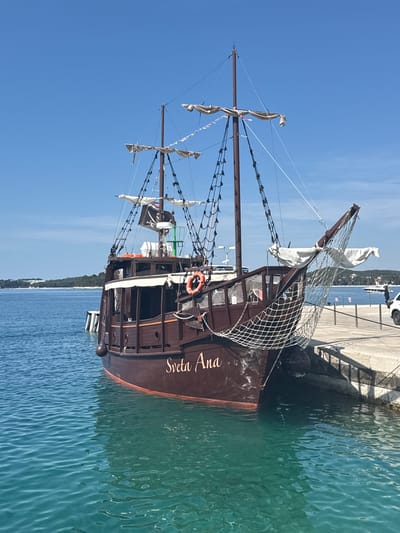 The image depicts a brown pirate ship named "Sveta Ana" docked at a concrete pier in Grad Rovinj, Croatia. The ship has multiple masts with sails, a pirate flag, and safety equipment such as a life ring. The water around the ship is clear and blue-green. The sky is a clear blue. A strip of land with trees can be seen in the background across the water. A car is parked on the pier further down.