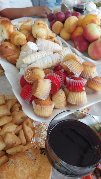 In Novonikolaevka, Kyrgyzstan, a close-up, eye-level shot captures a table laden with a variety of baked goods and fruit, with a glass of dark liquid in the foreground. The baked goods include croissants, pastry horns filled with cream and dusted with powdered sugar, and mini muffins served in red and white paper liners. Some muffins are sprinkled with poppy seeds. To the left, a pile of fried dough pieces, resembling small pillows, are visible. A partially visible piece of flatbread with sesame seeds sits in the lower left corner. Adjacent to the baked goods, a cluster of red plums and yellow and red peaches are arranged on a white platter. In the bottom right corner, a glass filled with a dark, viscous liquid, possibly juice or wine, is positioned on a light-colored tablecloth. A person's hands, one with a ring, are partially visible in the upper left background, suggesting a meal or gathering is taking place. The lighting is natural and diffused, casting soft shadows.