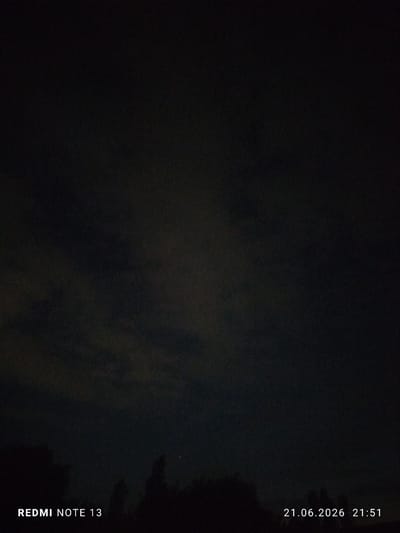 This media file is a nighttime photograph taken in Асан-Чек, Kyrgyzstan. The scene is dominated by a dark sky with subtle cloud formations, suggesting it is nighttime. In the lower portion of the image, a silhouetted treeline is visible against the dark sky, indicating a natural or rural setting. A single, faint light source, possibly a star or a distant light, is present in the sky above the treeline.

The bottom left corner of the image contains the visible text "REDMI NOTE 13", which is likely a watermark indicating the device used for capture. In the bottom right corner, the date and time are displayed as "21.06.2026 21:51". This suggests the photograph was taken on June 21, 2026, at 9:51 PM. The overall mood is dark and quiet, characteristic of a night scene. There are no people or prominent objects visible, with the focus being on the sky and the treeline. The weather appears to be partly cloudy or clear enough to allow for some visibility of the sky.