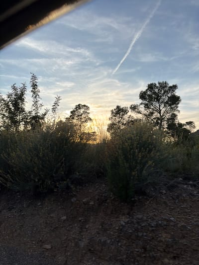 A landscape view, potentially within the Urbanització El Totxar area of Spain, with the sun low on the horizon, creating a sunset or sunrise effect. The sky is streaked with clouds and a visible contrail. Silhouetted trees and shrubs dominate the landscape against the bright sky. The foreground consists of a dirt or gravel ground surface. The image appears to be taken at ground level, with a metallic object visible in the lower left portion of the frame. No people are visible in the scene.