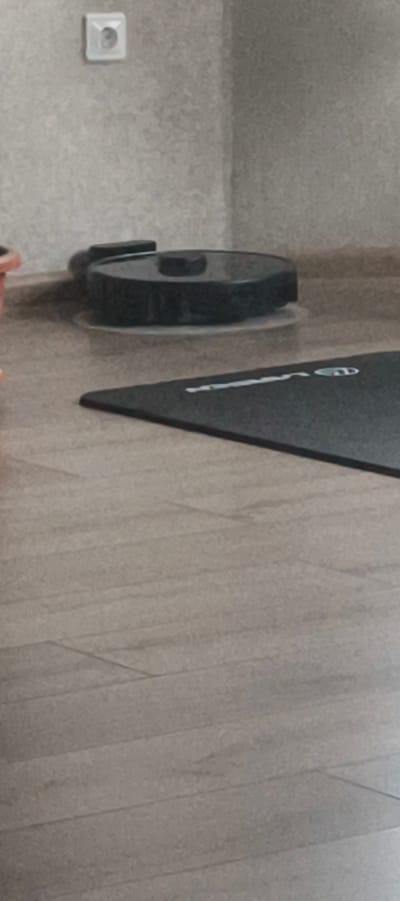 This media file features a low-angle shot of a domestic interior. The primary objects in focus are a black robotic vacuum cleaner positioned on a light-colored floor, and a black yoga mat with a subtle white logo or marking. In the background, a white electrical outlet is visible on a textured, light gray wall. The lighting suggests an indoor setting, likely during the daytime, with no strong indicators of weather conditions. The scene appears to be a quiet, possibly tidy, living space or a room dedicated to exercise. There are no people visible, and the overall mood is neutral. The location context indicates Aktanyshbash, Russia. The visible text on the yoga mat is too indistinct to read clearly.