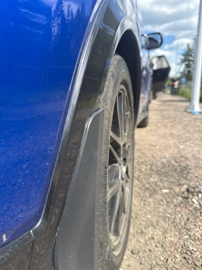 An extreme close-up shot focuses on the rear wheel and fender of a blue car, viewed from a low angle. The car's blue paint has reflections of trees. A black plastic fender flare is visible, with a mud flap attached below it. The wheel features a multi-spoke silver rim with a tire that has some visible lettering. The background is out of focus, showing a gravel path, a sidewalk with a white pole, and a second car with its door open. In the distance, a person in dark clothing walks away from the camera. The scene suggests an outdoor location, possibly a parking area or a road shoulder, with hints of a suburban or semi-rural environment given the presence of trees and a natural landscape. The specific location in Серебряный Бор, Russia, is implied by the context but not visually confirmed within the image itself.