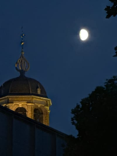 The image shows a nighttime view of a church tower with a dome and a cross on top, illuminated by artificial light. A partially illuminated moon is visible in the dark blue sky. Silhouetted trees are in the foreground and to the right. The church tower has architectural details such as arched windows and ornate decorations. The overall scene suggests a peaceful, serene evening in a city. This is the Westerkerk in Deventer, Netherlands.