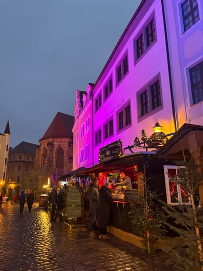 This image depicts an evening scene in Meissen, Germany, likely a Christmas market. The sky is a deep grey-blue, characteristic of dusk or early night. The cobblestone street, damp from recent rain, reflects the various lights, adding to the atmosphere.
On the right, a prominent multi-story building with a pale facade is illuminated by striking purple and pink lights, giving it a surreal glow. Numerous windows are visible across its surface. Along its side, the text "ALBRECHTSBURGER MEISSEN" is clearly legible, providing a precise location cue. An ornate, old-fashioned lantern hangs from this building.
To the left, several wooden market stalls line the street. These stalls are festive, adorned with evergreen branches and strings of warm, twinkling fairy lights. One stall, dark-painted with a white-framed window, shows red curtains and internal lights. Signs on the stalls advertise typical German market fare: "Glühwein," "Punsch," "Eierpunsch," "Heißer Hirsch," "Met," "Kinderpunsch," "Bratwurst," "Waffeln," and "Kaffee," with prices such as "€ 3.50" indicating sales in Euros.
Several people are visible in the scene, walking along the street and congregating around the market stalls, some appearing to interact with vendors. Their winter clothing suggests cold weather. In the background, other historical buildings with traditional pitched roofs can be seen, hinting at the town's architectural character. The overall scene conveys a festive and cozy ambiance, typical of a European winter market.