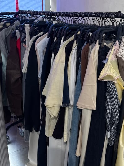 The image shows a close-up view of a clothing rack filled with various garments. The rack is made of black metal, and the clothes are hung on black plastic hangers. The clothing items vary in color and style, including what appear to be shirts, dresses, and possibly jackets. Some of the visible colors are brown, gray, white, black, blue, and yellow. The background is blurred, but suggests an indoor setting, possibly a store or a closet, with some lighting visible on the floor. There are no people visible in the image. The scene is consistent with a retail environment where clothing is displayed for sale. It is not possible to determine the specific location within Родниковское сельское поселение, Ukraine, from this image alone, as it lacks any identifying landmarks or signage.