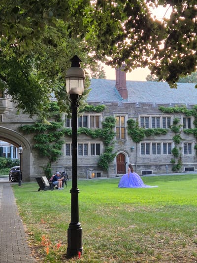 The image depicts a scene on the campus of Princeton University in Princeton, New Jersey. In the foreground, a black lamppost stands on a grassy area. To the left, a paved walkway runs parallel to the grass, with a cobblestone path adjacent to it. An arched entryway to a building is visible on the far left. In the middle ground, two individuals are seated on dark-colored benches on the grass, facing away from the camera and towards a building. One person is holding a phone and the other is looking at it. Behind them, a bicycle is leaning against a bench. A person in a voluminous, light purple ball gown stands in the grass, facing away from the camera, with their back to a large stone building that features many windows and is covered in ivy. The building has a slate roof and a brick chimney. The scene is illuminated by the soft light of late afternoon or early evening, with sunlight filtering through the leaves of trees that overhang the area.