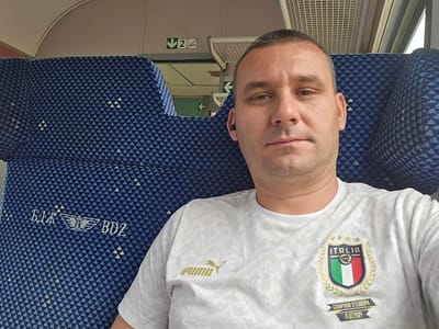 A middle-aged man with short hair is photographed from an upside-down perspective, lying on a blue seat with a triangular pattern. The seat also displays the text "BJA BDZ," potentially indicating a transportation company. The man is wearing a white t-shirt with the Italian national football team's logo and "Puma" branding. The logo includes the words "ITALIA," "CAMPIONI D'EUROPA," and the date "11.07.2021." The background features a train interior with a white wall, an air vent, and signs with arrows. This is unlikely to be in Trastikovo, Bulgaria. It is more likely this is an interior train seat in Italy.