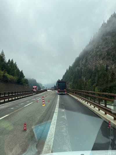 The image depicts a multi-lane highway near Brenner - Brennero, Italy. Traffic is congested, with several trucks visible in the lanes ahead. The weather is overcast, with low-hanging clouds or fog partially obscuring the mountains flanking the highway. Construction or roadwork is indicated by the presence of red and white striped barriers along the shoulder. The view is from inside a vehicle, with the interior and the vehicle's side window partially visible on the right side of the image.