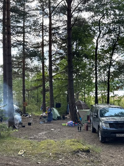 A group of people are camping in a forest in Portovoe, Russia. The scene shows a campsite with tents, a campfire, and a car. A woman in a blue dress is standing at a table, preparing food. Smoke is rising from the campfire, and clothes are hanging on a line. The ground is covered with pine needles and dirt. In the background, there are tall trees and a body of water.