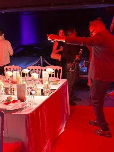 The image shows a man in a black suit taking a picture with a camera at a table set with white linens and white roses. The man is wearing a black suit jacket and black pants. He is holding a camera in his right hand and his left arm is extended towards the camera, as if he is taking a picture. There are three other people in the background. There is a partially visible white car, a dark car, and a red carpet. The table is set with white plates, white napkins, silver utensils, glasses of water, and two white roses in vases. The environment is dimly lit with a red and purple lighting scheme. The location is Beausoleil, France. The coordinates are 43.749 latitude and 7.429 longitude.